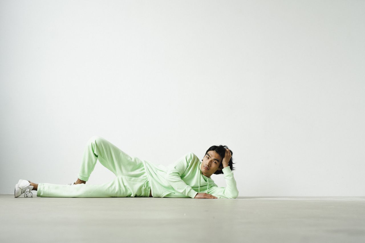 A man in a green tracksuit lies on the floor with hand on head, against a white background.